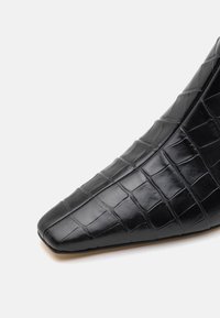 Bottine en cuir noir avec un bout pointu, présentant un motif crocodile texturé et une surface lisse.