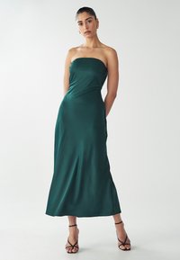 Abito in satin verde senza spalline con un corpetto aderente e una gonna fluente, abbinato a sandali neri con tacco alto e lacci.
