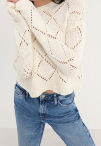 Gebreide trui in crème, met open ruitpatronen en ribbels aan de mouwen, gecombineerd met hoge taille blauwe jeans. Verkorte snit.