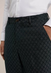 Detailní záběr na tmavě vzorované kalhoty s hnědým knoflíkem, které jsou nošeny s bílou košilí, s jednou rukou v kapse.