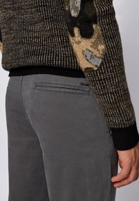 Stickad tröja i mörkgrönt och beige med ett strukturerat mönster, kombinerad med figurnära grå byxor med bakfickor och subtila sömnadsdetaljer.