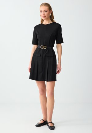 Vrouw draagt een zwarte jurk met korte mouwen en een gouden gesp riem, zwarte platte schoenen en gouden oorbellen tegen een effen lichte achtergrond.