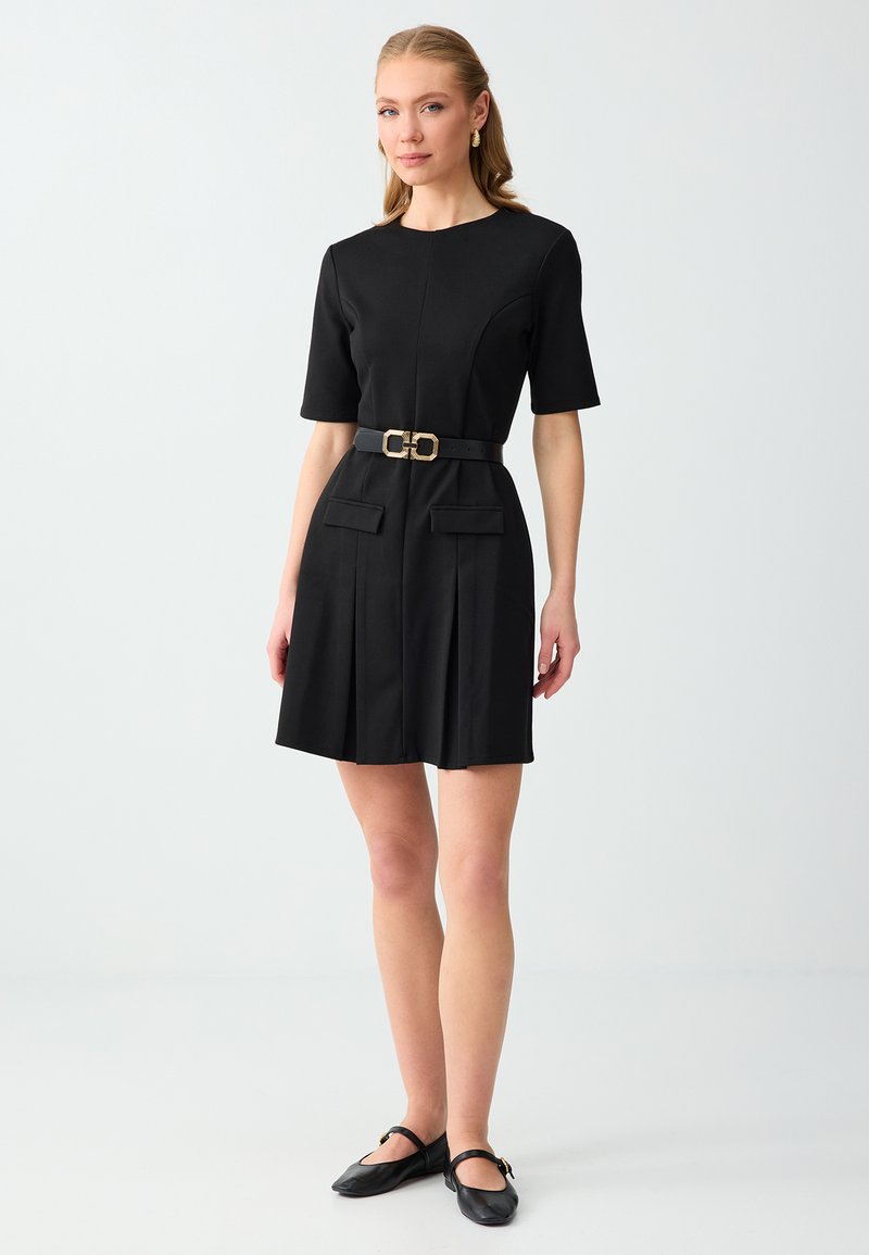 Femme portant une robe noire à manches courtes avec une ceinture à boucle dorée, des ballerines noires et des boucles d’oreilles dorées sur un fond clair uni.