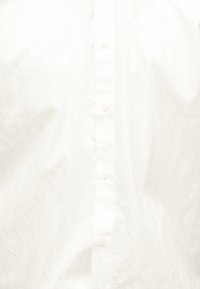 Μπροστινή όψη λευκού πουκαμίσου με κουμπιά ορατά κατά μήκος της λωρίδας, εμφανίζοντας διακριτικά ζάρια και ένα αχνό καπιτονέ μοτίβο στη αριστερή πλευρά.