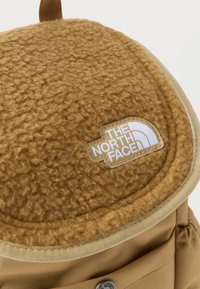 Καφέ φλις κάλυμμα και τμήμα υφασμάτινου σακιδίου με λευκό κεντημένο λογότυπο The North Face στο κάλυμμα.