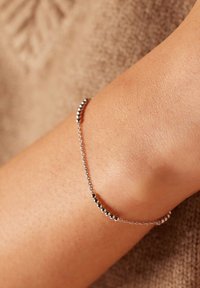 Zilveren schakelarmband met kleine ronde kralen, gepositioneerd op gebruinde huid, tegen een zachte, gestructureerde bruine achtergrond.