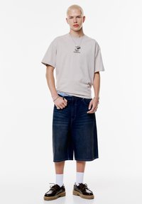 Beige kortærmet t-shirt med "HONDA" logo, parret med mørkeblå denim shorts og sorte sko med tykke såler, båret med hvide sokker.