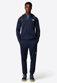Marinblå zip-hoodie och matchande joggers i mjukt tyg. Har svarta axelaccenter och The North Face-logotyp i vitt.