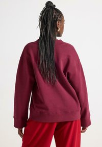Bordeaux oversized sweatshirt med ribbad halsringning och muddar, ihop med röda byxor. Strukturerad tyg, avslappnad passform och sänkta axlar.