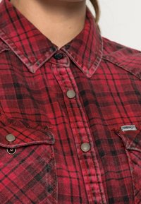 Camisa de cuadros rojos con cuello abotonado, botones grises y bolsillos parche. La tela tiene un aspecto suave y texturizado con un patrón a cuadros.