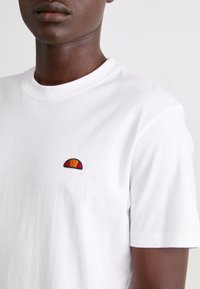 Weißes Baumwoll-T-Shirt mit einem kleinen, gestickten Halbkreis-Logo in Rot, Orange und Schwarz. Es hat einen klassischen Rundhalsausschnitt und kurze Ärmel.