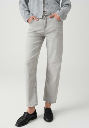 Personne portant un jean droit gris clair, un cardigan en tricot gris et des chaussures noires sans lacets, debout les mains dans les poches.
