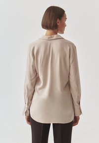 Beige long-sleeve shirt made of smooth fabric, features a classic collar, side slits, and buttoned cuffs, shown from the back.