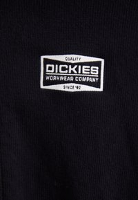 Zwarte stof met een rechthoekig label waarop de tekst "QUALITY DICKIES WORKWEAR COMPANY SINCE '92" staat in witte en gouden accenten.