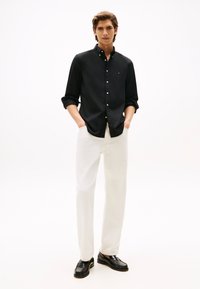 Noormees, pruunide juustega, kes kannab musta nööbitavat särk, valgeid pükse ja musti mokasiine, seisab käed taskutes valge tausta ees.