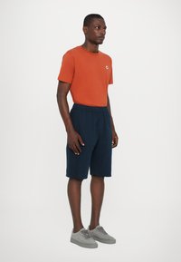 T-shirt en coton orange avec un col rond, shorts bleu marine à taille élastique et sneakers en suède gris. Design simple avec une coupe décontractée.