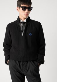 Felpa nera a maglia con colletto alto e zip a metà, tessuto testurizzato, logo circolare blu sul petto, abbinata a pantaloni neri e occhiali da sole.