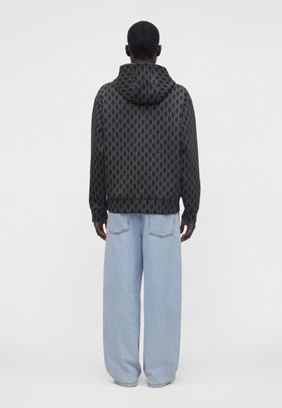 Homme vu de dos portant une veste à capuche noire à motifs et un jean large bleu clair sur un fond clair uni.