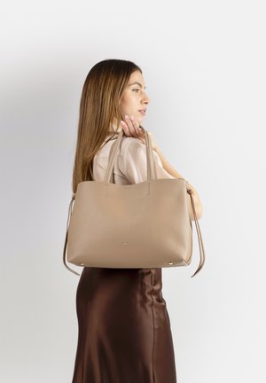 Giovane donna con lunghi capelli castani che porta una grande borsa a tracolla beige, indossa una camicetta chiara e una gonna di raso marrone su sfondo bianco.