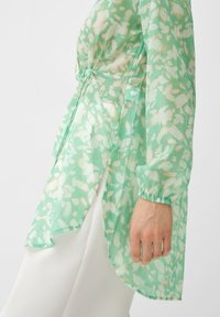 Donna che indossa una blusa trasparente floreale verde e bianca con vita arricciata e pantaloni bianchi, mano rilassata al fianco, sfondo chiaro.