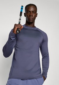Langærmet sports T-shirt i mørkegrå stof med slidse overflade og rund hals. Modellen holder et sort og blåt tennisracket.