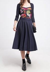 Il vestito presenta un corpetto a fantasia floreale con una gonna svasata di un blu navy scuro. L'outfit è completato da un cardigan aderente nero e stivaletti neri alla caviglia.
