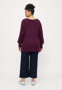 Langärmeliger, gerippter lila Pullover mit weitem Ausschnitt, kombiniert mit dunkelblauen weiten Jeans und silbernen Ballerinas.