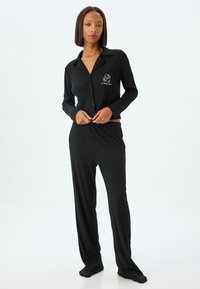 Conjunto de pijama negro con mangas largas y pantalones. Presenta un cuello, cierre de botones y un estampado gráfico en la parte izquierda del pecho. Textura suave.