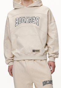 Felpa color crema con cappuccio, dotata di tasca a marsupio frontale, vestibilità oversize e testo stampato nero. Polsini e orlo a costine, realizzata in materiale morbido.