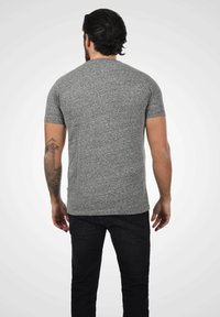 Camiseta gris de manga corta hecha de tejido suave, con cuello redondo y una silueta ajustada, combinada con jeans oscuros y un tatuaje visible en el brazo.