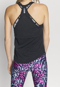 Svart träningslinne med en texturerad design, som har korslagda axelremmar och logodetalj. Kombinerat med livfulla, mönstrade tights.