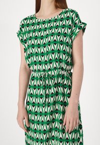 Grün- und cremefarbenes, gemustertes Kleid mit einem geometrischen Design, das schwarze Akzente aufweist. Es hat kurze Ärmel, einen runden Ausschnitt und eine verstellbare Kordelzug-Taille.