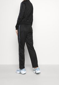 Svart träningsjacka och byxor med sidostripes som har ett logomönster. Texturerad tyg och åtsittande design. Ljust blå sneakers.