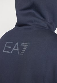 Felpa blu navy con una texture morbida, con il logo riflettente EA7 sulla parte superiore della schiena e un design casual e rilassato.