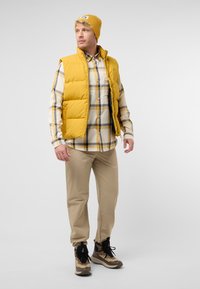 Gilet imbottito giallo sopra una camicia a quadri, pantaloni beige e stivali marroni mimetici. Dettagli evidenti includono un berretto con logo e polsini con bottoni.