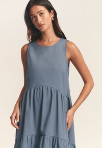 Šviesiai mėlyna, be rankovių suknelė su apvaliu iškirpimu, sluoksniuotu sijonu ir lygiu audiniu, turinti subtilią tekstūrą ir be matomų raštų.