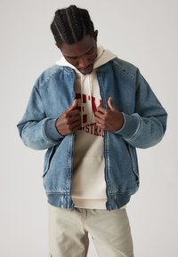 Chaqueta bomber de mezclilla en azul claro con puños y dobladillo acanalados. Llevada sobre una sudadera crema con letras rojas en negrita. Diseño minimalista.