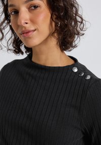 Kvinde med krøllet hår iført en sort ribbet top med tre sølvknapper på skulderen, mod en ensfarvet baggrund.