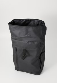 Sac à dos en nylon noir avec une forme rectangulaire haute, un dessus zippé et une poche latérale. Comprend une bandoulière ajustable et des détails en métal à l'extérieur.