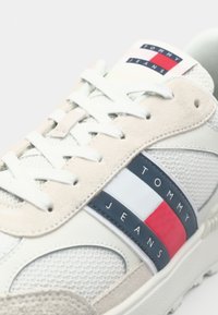 Bijela i bež tenisica s mrežastim i antilop materijalom, okrugli vrh, kontrastne mornarskoplave i crvene pruge s "TOMMY JEANS" logotipom.