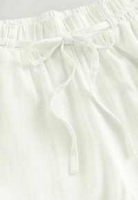 A fehér vászon sort elasztikus derékkal és kötőfűzővel rendelkezik. A szövet puha, könnyű tapintású, és enyhén áttetsző megjelenésű.