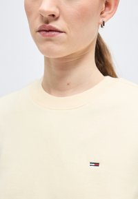 Γυναίκα με κρεμ πουλόβερ με στρογγυλή λαιμόκοψη, με μικρό κεντημένο λογότυπο σε κόκκινο, άσπρο και μπλε στο στήθος και ασημένιο κρίκο στο αριστερό αυτί.