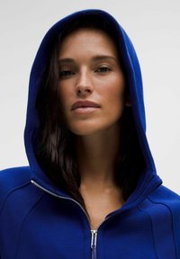 Felpa con zip blu, dotata di cappuccio, caratterizzata da una texture liscia e una zip argentata. Il tessuto è morbido e aderente, con dettagli di cucitura sulle spalle.