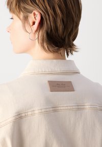 Beige denimjacka med en läderpatch på ryggen med präglad logotyp. Synliga sömnadsdetaljer längs sömmarna.