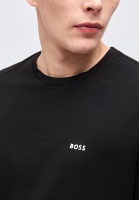 Κοντινό πλάνο ενός ατόμου που φοράει μαύρο πουκάμισο με τη λέξη "BOSS" τυπωμένη με λευκά γράμματα στο στήθος, στραμμένο ελαφρώς προς τα δεξιά.