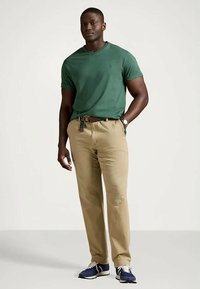 Άνδρας που φορά πράσινο κοντομάνικο μπλουζάκι, μπεζ παντελόνι με υφασμάτινη ζώνη, μπλε αθλητικά παπούτσια, στέκεται μπροστά σε απλό ανοιχτό φόντο.