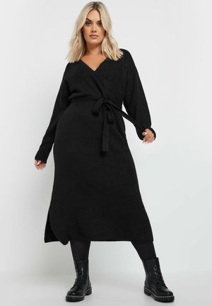Schwarzes Wickelkleid mit langen Ärmeln, V-Ausschnitt und seitlichem Schlitz. Akzentuiert mit einem passenden Gürtel. Gestylt mit schwarzen Schnürstiefeln.