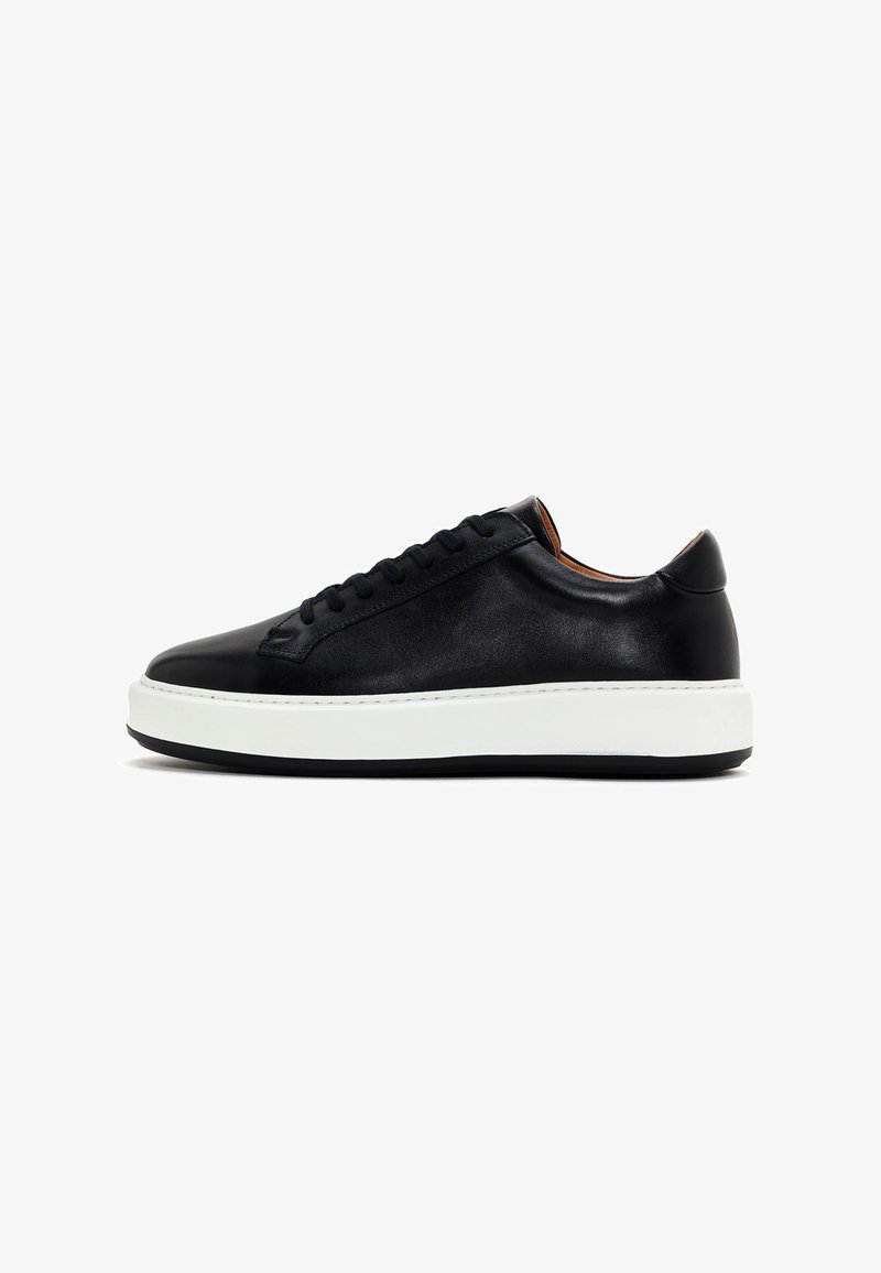 Zapatilla de cuero negro con punta redondeada, suela de goma blanca plana y cordones negros. Textura suave y diseño minimalista.