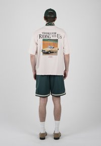 Personne portant un T-shirt rose pâle avec une voiture vintage et le texte « Thanks for Riding With Us », short vert foncé, chaussettes blanches, chaussures beige et casquette noire.