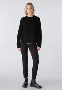 Schwarzer gestrickter Pullover mit gerippter Textur, seitlichen Schlitzen und Rundhalsausschnitt. Kombiniert mit schwarzen Hosen und glänzenden schwarzen Slippern.